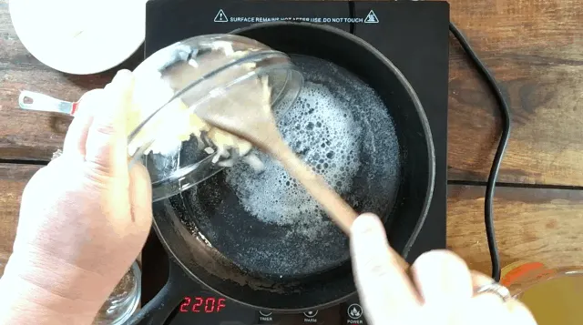 a black frying pan with butter and oil melted in it with a bowl of chopped onions being poured into it from glass bowl with wooden spoon
