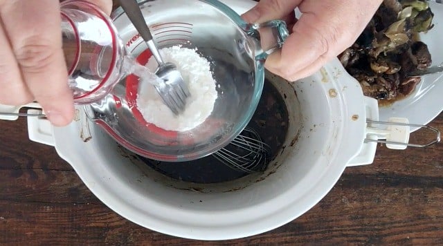 hand pouring water from small glass into glass fluid measure with cornstarch in bottom. Crockpot and plate of meat in background