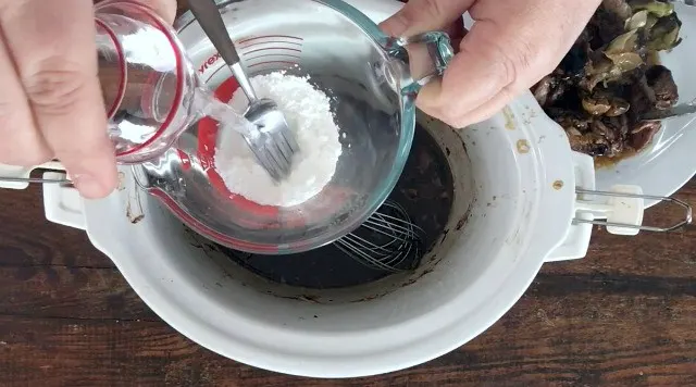 hand pouring water from small glass into glass fluid measure with cornstarch in bottom. Crockpot and plate of meat in background