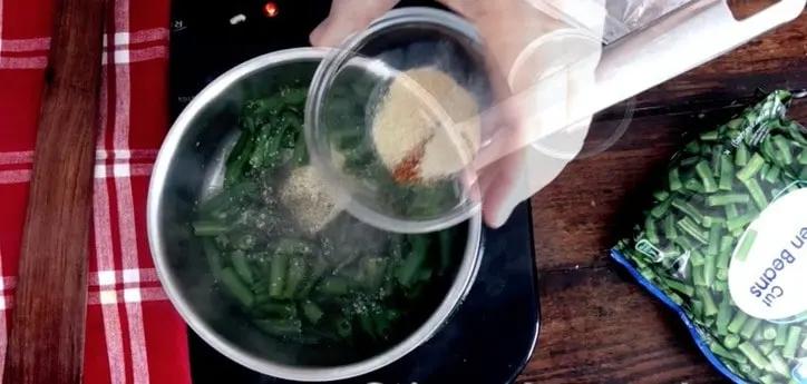 A small glass bowl adding seasonings to saucepan of green beans cooking below