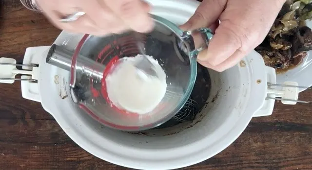 hand using fork to mix cornstarch slurry in a glass fluid measure over the top of a crockpot. Plate of meat in the background