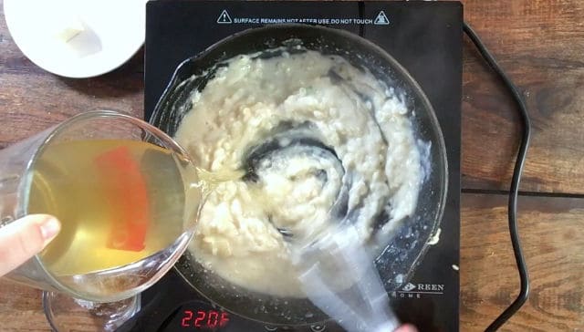 A whisk stirring the mixture in the frying pan while a liquid measure pours stock into the pan for how to make gravy without drippings