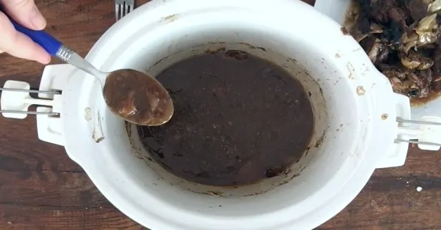 a spoon with blue handle holding the cornstarch gravy over the crockpot of gravy with plate of meat in the background