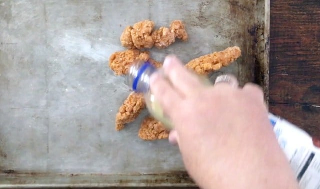 hand adding oil to chicken strips on a baking pan