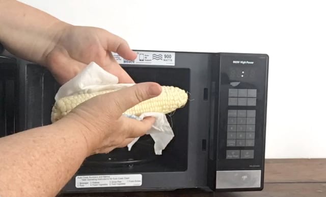 two hands holding corn on the cob wrapped in paper towel microwave in background