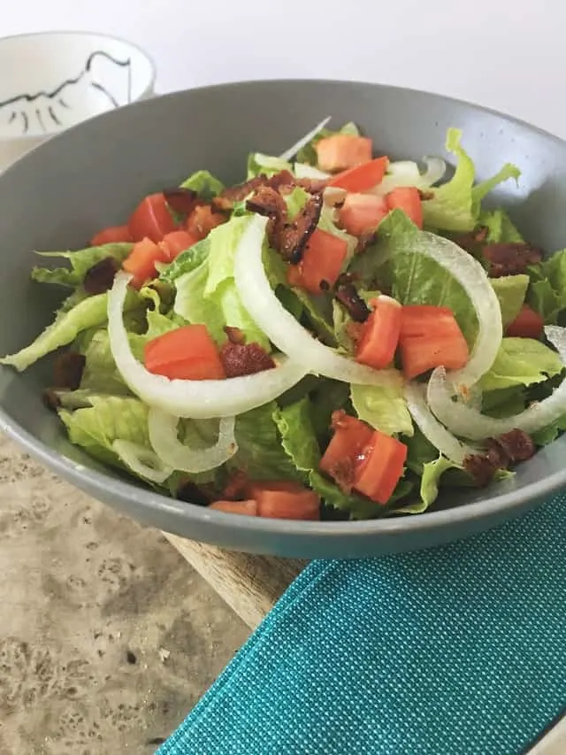 side view of gray bowl of greens, onion, tomato and bacon with dressing on brown table