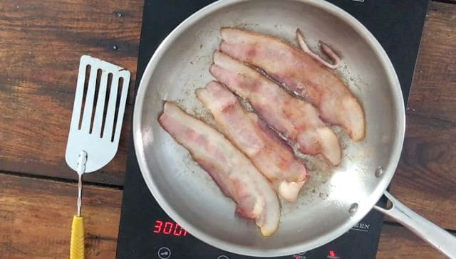 four slices of bacon frying in a frying pan with spatula
