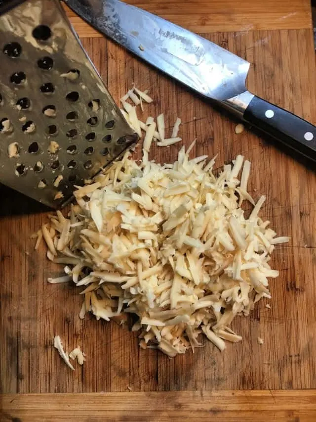 grated white cheese on cutting board with grater and knife