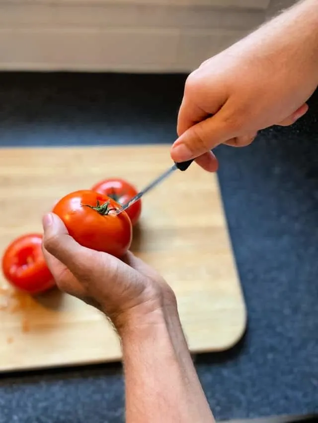 hand holding tomato and another hand poking a paring knife next to the stem