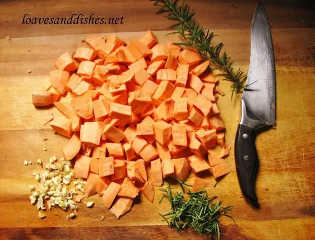 A chopped sweet potato on a cutting board with garlic and rosemary