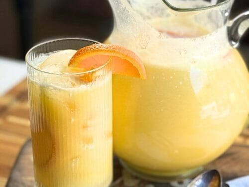Glass pitcher and tumbler of Orange Coconut Refresher with an orange slice garnish, ice cubes, and a spoon on a wooden serving board.