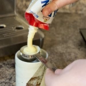A hand pouring sweetened condensed milk from a can into a cup.