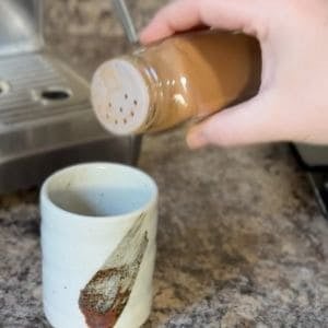 A hand sprinkling ground cinnamon into a cup with sweetened condensed milk