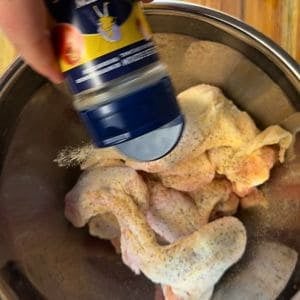 hand sprinkling Mortons Nature Seasoning on top of raw chicken wings in a bowl