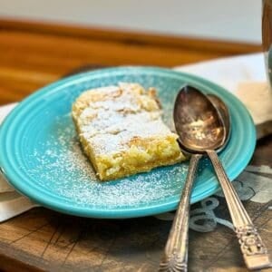 gooey butter cake on a blue plate with powdered sugar sprinkled on top and two spoons
