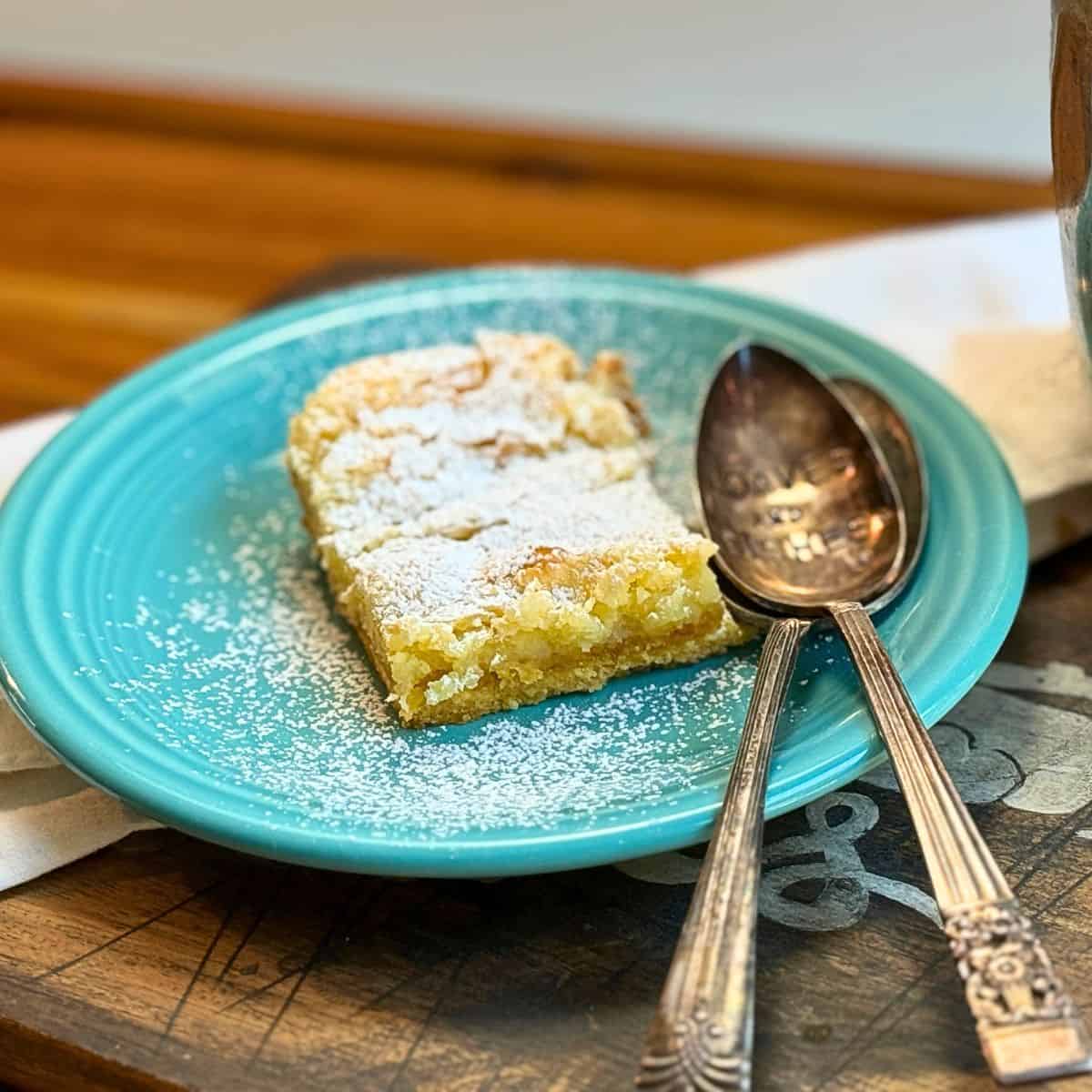gooey butter cake on a blue plate with powdered sugar sprinkled on top and two spoons