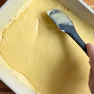 spatula spreading cream cheese mixture over the cake mixture in a baking dish