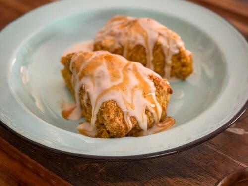 Starbucks Copycat Pumpkin Scones Two golden pumpkin scones covered with vanilla icing and pumpkin spice drizzle on a blue plate.