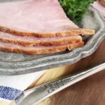 The edge of three pieces of ham on a gray plate with a napkin and the handle of a fork